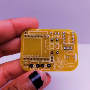 Close-up of a hand holding a small yellow custom PCB labeled 'DMB V2', designed for an ATtiny85 and DFPlayer Mini. The board includes silkscreen labels for components like a speaker, reed switch, resistors, and capacitor.