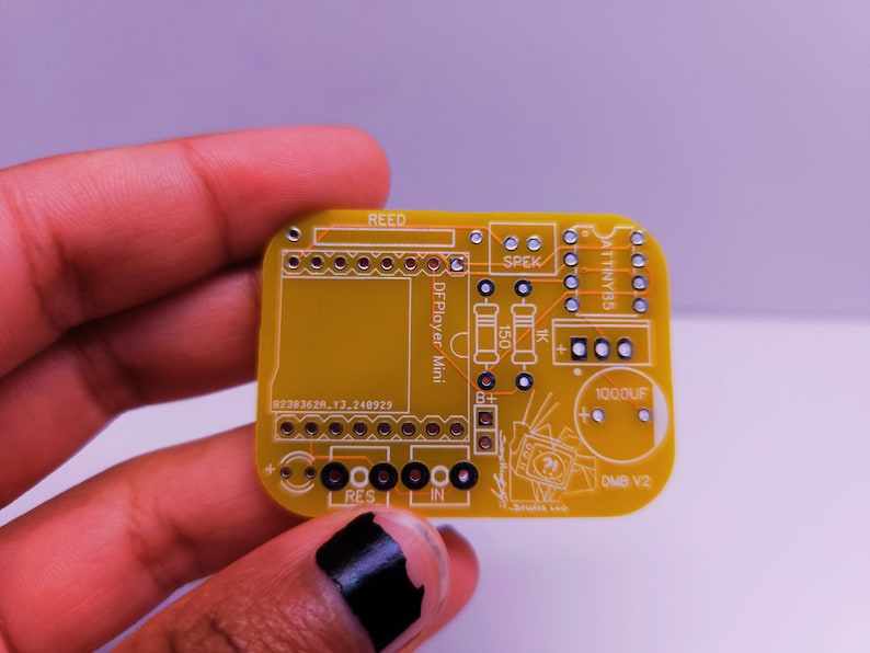 Close-up of a hand holding a small yellow custom PCB labeled 'DMB V2', designed for an ATtiny85 and DFPlayer Mini. The board includes silkscreen labels for components like a speaker, reed switch, resistors, and capacitor.