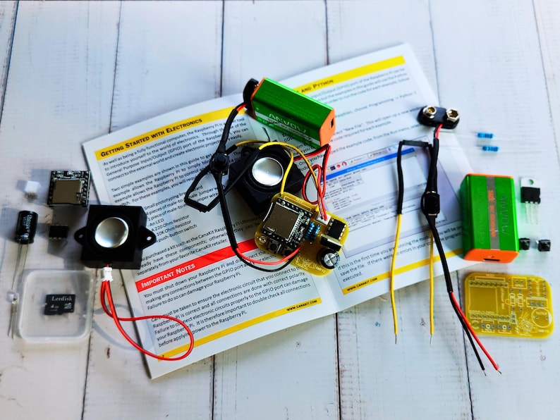 Flat lay of electronics components arranged around an open instruction manual titled 'Getting Started with Electronics.' Items include speakers, a microcontroller board, capacitors, resistors, a microSD card, battery clips, 9V batteries, jumper wires, and a custom yellow PCB.