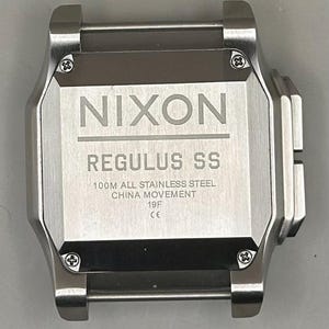 K&ouml;nnte beinhalten: Eine Edelstahl-Nixon Regulus SS-Uhr. Das Zifferblatt der Uhr zeigt das Nixon-Logo und den Modellnamen. Der Text auf dem Zifferblatt weist darauf hin, dass es sich um 100M Edelstahl mit einem China-Uhrwerk handelt.