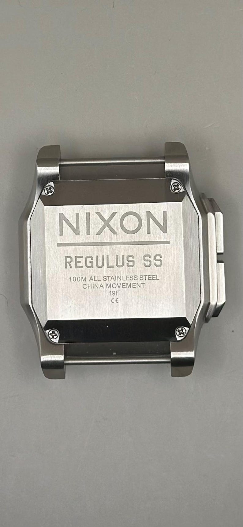 K&ouml;nnte beinhalten: Eine Edelstahl-Nixon Regulus SS-Uhr. Das Zifferblatt der Uhr zeigt das Nixon-Logo und den Modellnamen. Der Text auf dem Zifferblatt weist darauf hin, dass es sich um 100M Edelstahl mit einem China-Uhrwerk handelt.