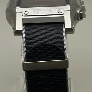 K&ouml;nnte beinhalten: Eine silberfarbene Digitaluhr mit einem schwarzen Nylonarmband. Das Zifferblatt ist rechteckig mit dem Markennamen "NIXON". Das Armband wird mit einer silberfarbenen Schnalle befestigt.