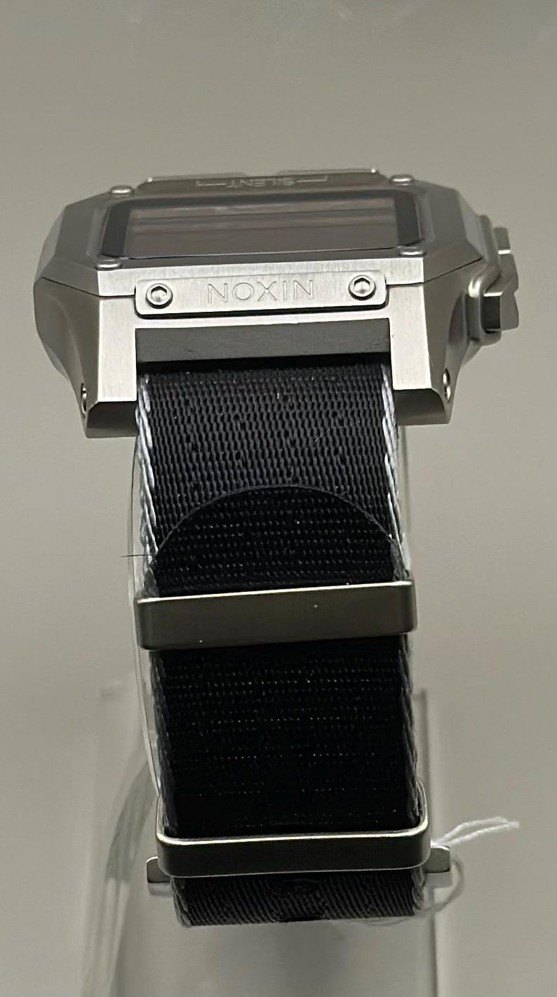 K&ouml;nnte beinhalten: Eine silberfarbene Digitaluhr mit einem schwarzen Nylonarmband. Das Zifferblatt ist rechteckig mit dem Markennamen "NIXON". Das Armband wird mit einer silberfarbenen Schnalle befestigt.