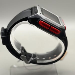 以下が含まれることがあります： シルバーの文字盤と赤いアクセントボタンが付いた黒いデジタル時計。時計は黒いラバーストラップで、透明なスタンドに展示されています。