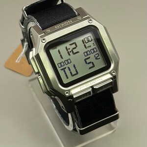 K&ouml;nnte beinhalten: Eine silberne Nixon Digitaluhr mit schwarzem Stoffarmband. Das Zifferblatt zeigt die Uhrzeit 11:12 Uhr an, mit zus&auml;tzlichen Digitalanzeigen. Das Armband ist schwarz und die Uhr wird auf einem transparenten St&auml;nder pr&auml;sentiert.