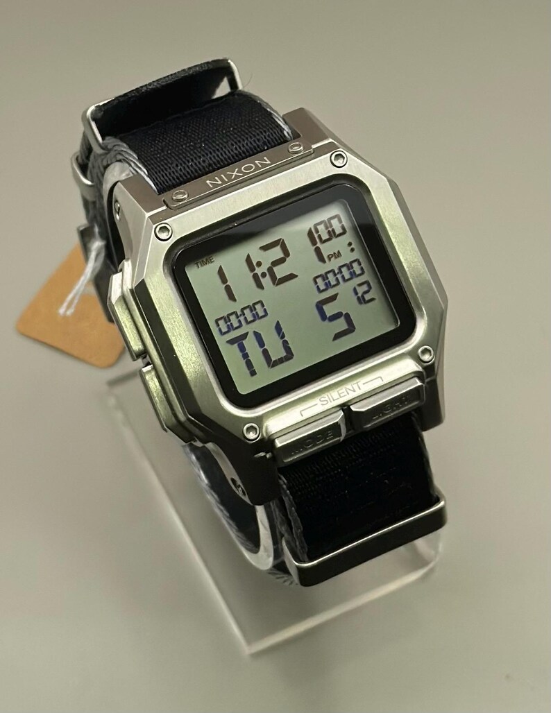 K&ouml;nnte beinhalten: Eine silberne Nixon Digitaluhr mit schwarzem Stoffarmband. Das Zifferblatt zeigt die Uhrzeit 11:12 Uhr an, mit zus&auml;tzlichen Digitalanzeigen. Das Armband ist schwarz und die Uhr wird auf einem transparenten St&auml;nder pr&auml;sentiert.