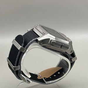 K&ouml;nnte beinhalten: Eine silberfarbene Digitaluhr mit einem schwarzen Stoffarmband. Das Zifferblatt der Uhr ist rechteckig, mit dem Markennamen "NIXON" sichtbar. Das Armband hat silberne Metallschlaufen. Die Uhr wird auf einem transparenten St&auml;nder pr&auml;sentiert.