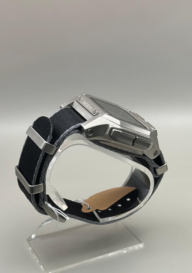 K&ouml;nnte beinhalten: Eine silberfarbene Digitaluhr mit einem schwarzen Stoffarmband. Das Zifferblatt der Uhr ist rechteckig, mit dem Markennamen "NIXON" sichtbar. Das Armband hat silberne Metallschlaufen. Die Uhr wird auf einem transparenten St&auml;nder pr&auml;sentiert.