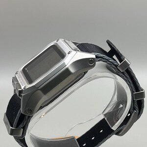 K&ouml;nnte beinhalten: Silberfarbene Digitaluhr mit schwarzem Stoffarmband. Die Uhr hat ein rechteckiges Zifferblatt und ein schwarzes Armband mit Metallakzenten. Die Uhr wird auf einem transparenten St&auml;nder vor neutralem Hintergrund pr&auml;sentiert.