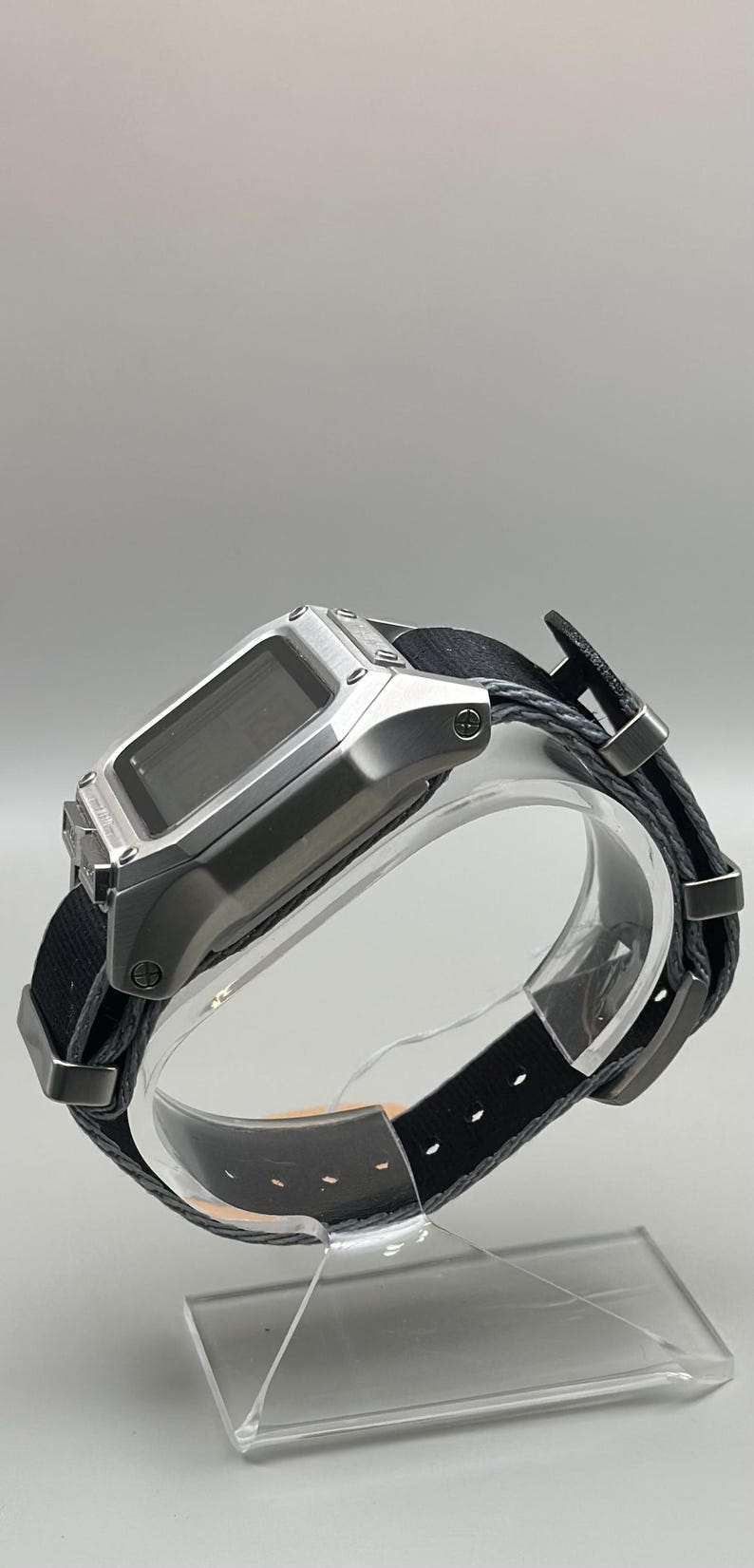 K&ouml;nnte beinhalten: Silberfarbene Digitaluhr mit schwarzem Stoffarmband. Die Uhr hat ein rechteckiges Zifferblatt und ein schwarzes Armband mit Metallakzenten. Die Uhr wird auf einem transparenten St&auml;nder vor neutralem Hintergrund pr&auml;sentiert.
