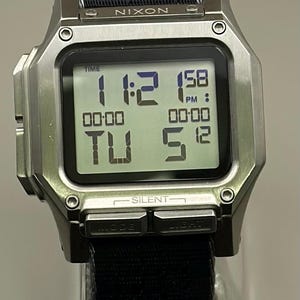 K&ouml;nnte beinhalten: Silberne Digitaluhr mit schwarzem Stoffarmband. Das Zifferblatt zeigt die Uhrzeit 23:12 Uhr sowie den Tag und das Datum an. Der Markenname "NIXON" ist auf der Uhr sichtbar.