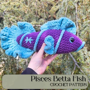 Puede incluir: Un pez Betta hecho a mano en crochet, en tonos morados y azules. El pez presenta un diseño detallado con rayas y aletas, y un símbolo en forma de estrella. El texto "Pisces Betta Fish Crochet Pattern" se muestra en la parte inferior.