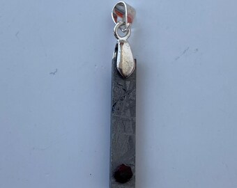 May include: A rectangular pendant with a silver bail and a dark, textured body. A small, dark red stone is set near the bottom. The pendant is approximately 1.5 inches long.