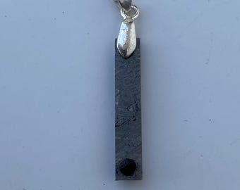 May include: A rectangular pendant with a silver bail and a dark gray, textured body. A small, dark red stone is set at the bottom. The pendant is designed to be worn as a necklace.