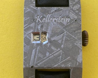 May include: A silver watch with a black leather band. The watch face is made of meteorite and has a digital display. The watch face also has the text "Kellerstein" engraved on it.