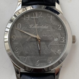 Könnte beinhalten: Eine silberne Armbanduhr mit einem strukturierten grauen Zifferblatt, mit dem Markennamen "Kellerstein". Die Uhr hat silberne Zeiger und Stundenmarkierungen, eine Datumsanzeige und ein schwarzes Lederarmband. Das Uhrengehäuse hat einen Durchmesser von ca. 3,8 cm.