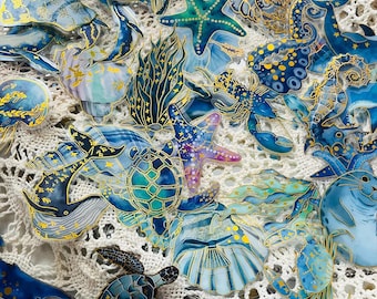 May include: A collection of ocean-themed stickers featuring various marine life, including whales, turtles, starfish, and seahorses. The stickers are primarily blue and gold, with intricate details and a glossy finish. The stickers are arranged on a white, textured surface.