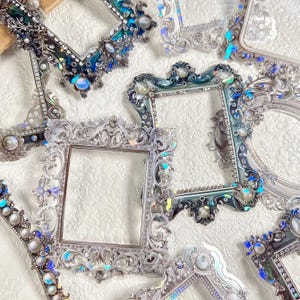 May include: An assortment of ornate picture frames in various shapes and sizes. The frames feature intricate designs with silver, blue, and iridescent accents, including pearls and crystals. The frames are arranged on a textured white surface.