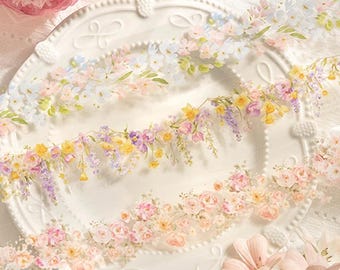 May include: Three rolls of decorative washi tape featuring delicate floral designs in shades of blue, yellow, and pink. The tape is displayed on a white plate with a beaded edge.