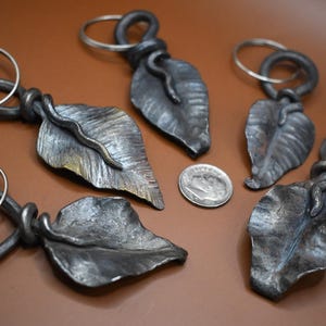 May include: Five handmade metal keychains shaped like leaves with a twisted metal ring. The keychains are a dark silver color.