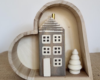 Può includere: Una scatola di legno a forma di cuore con una piccola casa di legno e un albero di legno bianco al suo interno. La casa ha un esterno dipinto di grigio e bianco con una porta e finestre bianche. L'albero ha una forma semplice e arrotondata.
