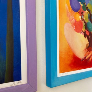 May include: Two framed art prints, one with a blue background and the signature "Ellie" and the other with a bright orange background and the text "We are held".