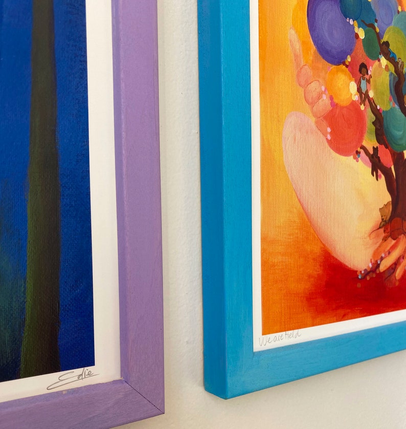 May include: Two framed art prints, one with a blue background and the signature "Ellie" and the other with a bright orange background and the text "We are held".