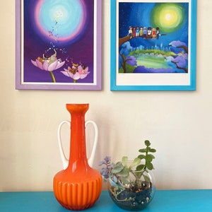 Puede incluir: Dos cuadros enmarcados con un jarr&oacute;n y un terrario de vidrio con suculentas sobre una mesa azul. La pintura de la izquierda presenta un fondo morado con un c&iacute;rculo blanco y una flor de loto rosa con un elefante gris y un mono marr&oacute;n. La pintura de la derecha presenta un fondo azul con una luna amarilla y un grupo de ni&ntilde;os sentados en una rama.