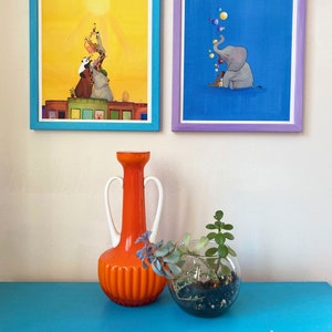Puede incluir: Dos ilustraciones enmarcadas, una con fondo amarillo y una pila de animales que alcanzan el sol, la otra con fondo azul y un elefante que sopla burbujas. Un jarr&oacute;n naranja y un terrario de vidrio con suculentas est&aacute;n sobre una mesa azul.