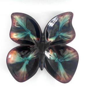Peut inclure: Un plat en céramique en forme de papillon avec une finition brillante. Le plat présente une couleur de base foncée avec des accents turquoise, marron et orange dans un motif unique. Les ailes sont divisées en sections.