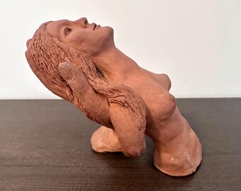 May include: A terracotta sculpture of a woman's torso and head, gazing upwards. The head is supported by a large hand. The sculpture is a reddish-brown color and appears to be made of clay.