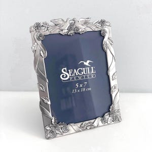 May include: A silver pewter picture frame with a floral and dove design. The rectangular frame features the text "SEAGULL PEWTER" and the dimensions 5 x 7 inches (13 x 18 cm).