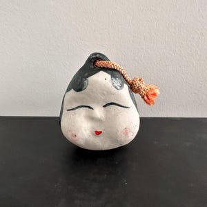 May include: A small ceramic figurine of a woman with a closed-eye smile. The figurine is painted white with black hair and red lips. It has a pink and orange cord attached to the top of its head.