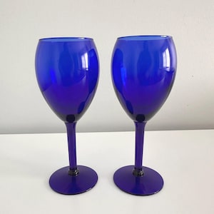 May include: Two cobalt blue wine glasses. Each glass has a rounded bowl, a slender stem, and a circular base. The glasses are made of a transparent material and reflect light.