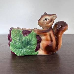 May include: A ceramic squirrel planter with a brown and tan body, holding a green leaf and purple grapes. The planter is designed to hold small plants or flowers. The squirrel has a detailed face and a bushy tail.