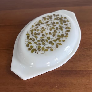 May include: White oval casserole dish with a green and gold berry design. The dish has a handle on each end and is made of glass. The dish is a vintage kitchenware item.