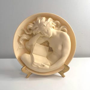 May include: A white ceramic wall plaque with a sculpted image of a woman reading a book. The plaque is on a gold metal stand.