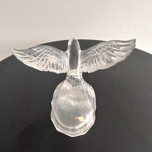 Puede incluir: Una escultura de cristal transparente de una paloma con las alas extendidas. La paloma est&aacute; posada sobre una base redonda.
