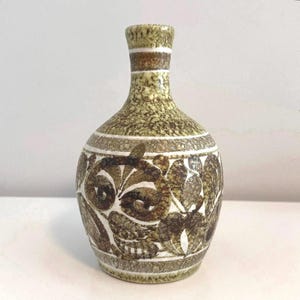 May include: A ceramic vase with a speckled green and brown finish. The vase features a bulbous body and a narrow neck, adorned with a repeating floral pattern in shades of brown and white. White bands encircle the neck and body.