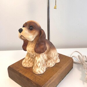 May include: A decorative dog-shaped lamp. The lamp features a ceramic dog figurine with brown and cream coloring, sitting on a wooden base. The lamp has a metal pole and a pull chain with a small gold-colored bell.