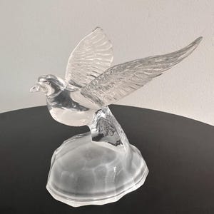 Puede incluir: Una figurita de paloma de vidrio transparente con las alas extendidas, posada sobre una base de vidrio esmerilado.