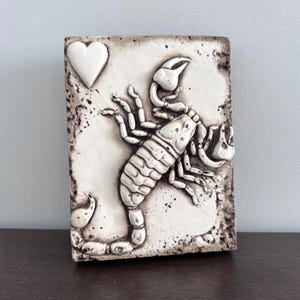 May include: A rectangular, off-white ceramic tile featuring a raised relief of a scorpion and a heart. The scorpion is detailed with segmented body and claws. The tile has a textured, aged appearance.
