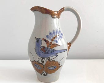May include: A small, white ceramic pitcher with a brown rim and handle. The pitcher is decorated with a blue bird perched on a branch with brown and blue flowers.