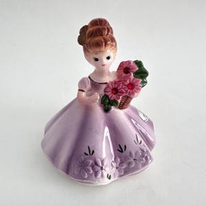 Peut inclure: Figurine en céramique d'une femme en robe lavande tenant un panier de fleurs roses. La figurine a des cheveux bruns et est ornée de détails floraux sur la robe. Une charmante pièce décorative.