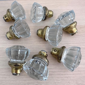 May include: Eight clear glass cabinet knobs with a faceted design. Each knob is mounted on a brass-coloured base. The knobs are arranged on a light-coloured wooden surface.