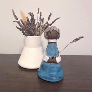 Peut inclure: Figurine en céramique d'une fille en robe bleue, tenant une brindille de lavande. La figurine a un visage brun et des cheveux gris. Un vase blanc avec de la lavande séchée et d'autres fleurs séchées est en arrière-plan.