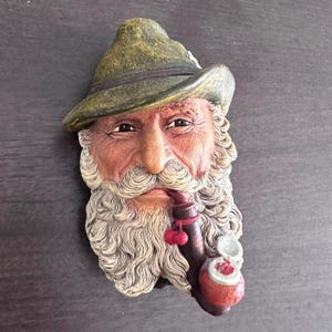 May include: A detailed, three-dimensional wall hanging depicting the face of an elderly man with a long, white beard and mustache. He wears a green hat and smokes a pipe with red cherries. The sculpture is painted in shades of brown, tan, and green.