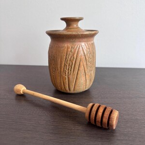 Peut inclure: Un pot à miel en céramique avec un couvercle et un bâtonnet à miel en bois. Le pot est de couleur marron clair avec un motif d'épis de blé. Le bâtonnet à miel est en bois clair et a une extrémité rainurée.