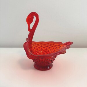 May include: A red glass swan shaped dish with a textured surface. The dish has a small base and a curved neck.