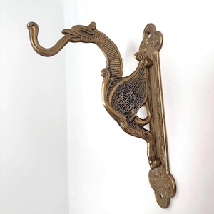 May include: A brass-coloured wall hook with a dragon design. The hook curves upwards from the dragon's head, with detailed wings and a decorative backplate. The dragon's wings have a textured, veined appearance.